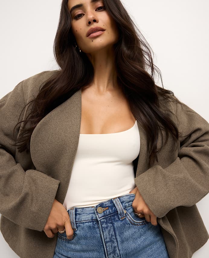 Model is wearing a brown wool coat over a white tank top and blue jeans.