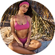 Model sits in the sand with her legs crossed in a pink headband and bikini.