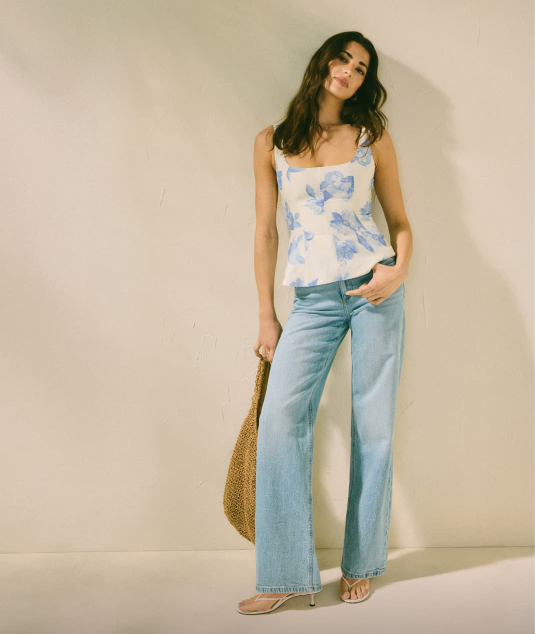 Model is wearing a white top with a blue floral print, light blue jeans, and a straw tote bag.