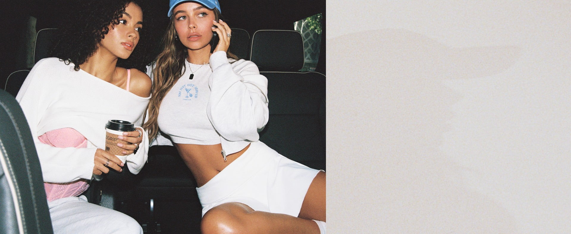 Two models sit in a car. One model is wearing an off the shoulder white sweater and white sweats. The other is wearing a cropped white graphic tee with a white hoodie and skirt.