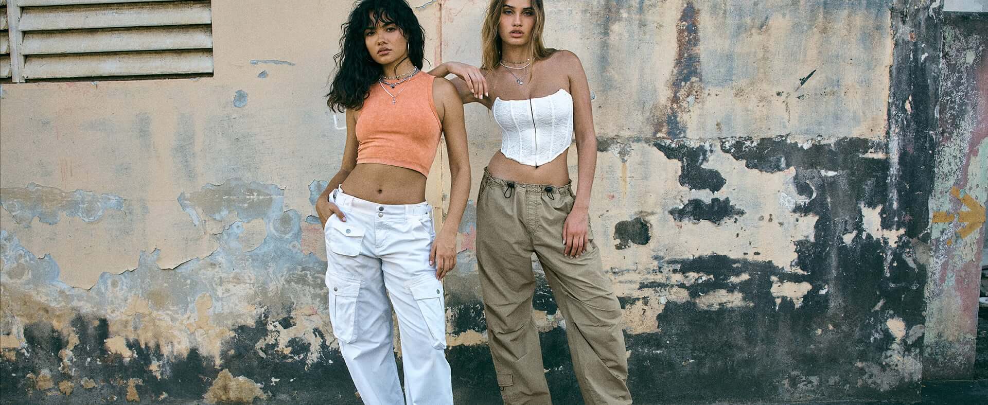 Two models pose on a sidewalk, one with an orange tank top and white cargo pants and the other in a white top with green cargos.