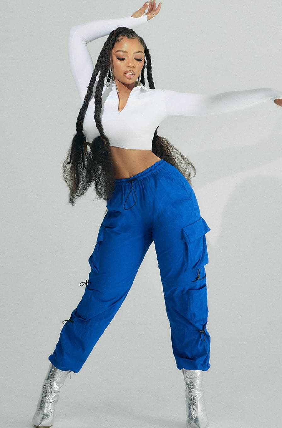 Musician Chloe Bailey wears a white zip up seamless top and blue parachute pants.