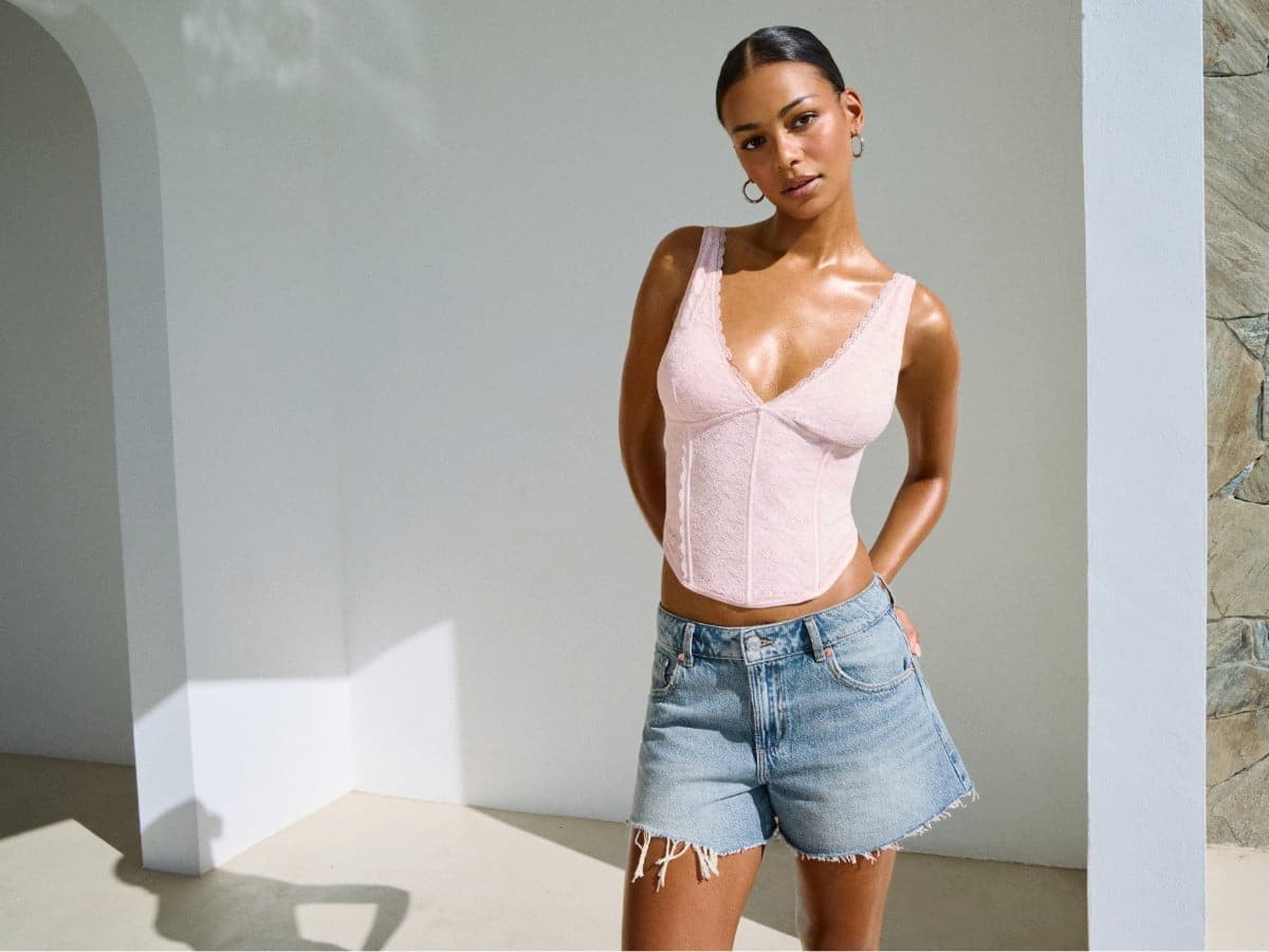 Model poses in a light pink tank top and distressed denim shorts. 