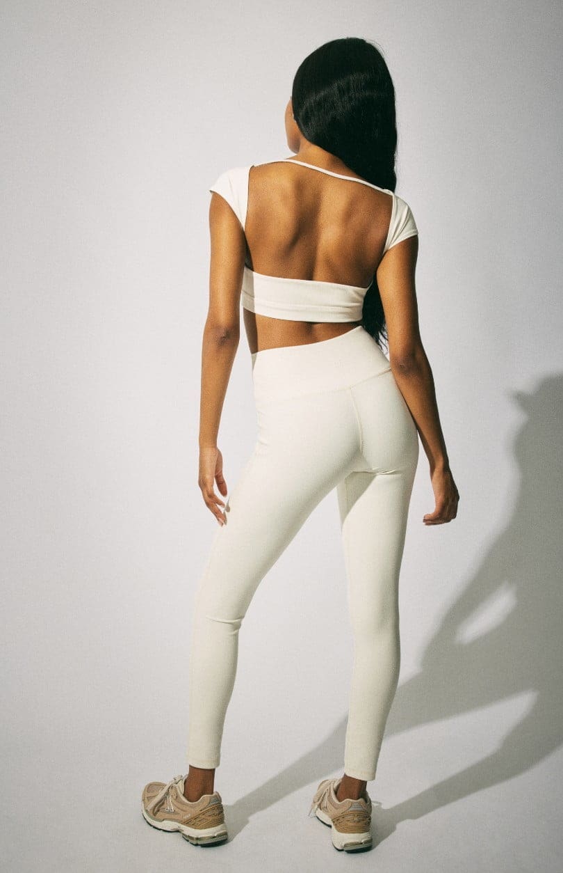 Model wears a cropped white tee with white leggings and a white sweater around her waist.