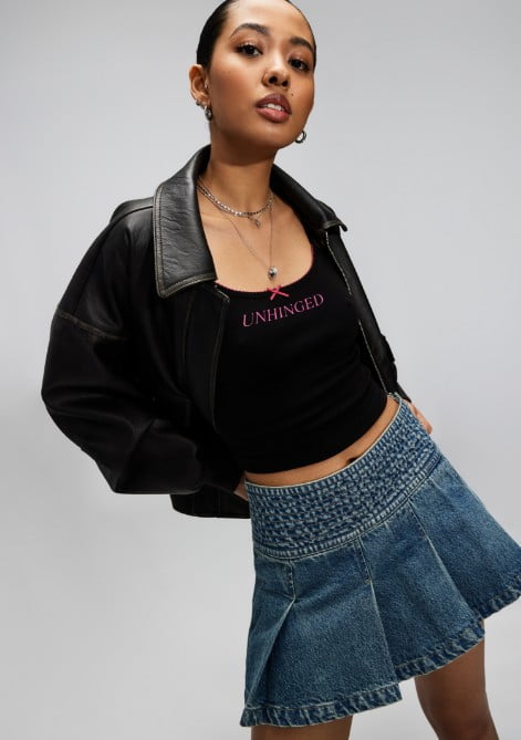 Model poses in a black  jacket over a black shirt with a pleated denim skirt and sneakers.