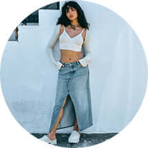 Model poses against a wall in a white tank top and shrug, denim shirt, and white clogs.